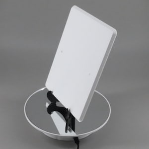 May include: A white rectangular product, possibly a tablet or display, is propped up on a reflective surface. The product is angled, revealing its side profile. The setup includes a black support structure and a white circular base.