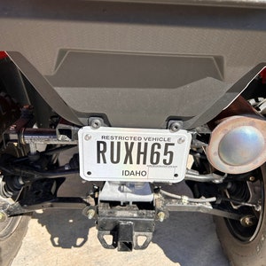 May include: A silver license plate with the text "RESTRICTED VEHICLE" and the license plate number "RUXH65" with the state "IDAHO" printed below. The license plate is attached to a black metal frame.