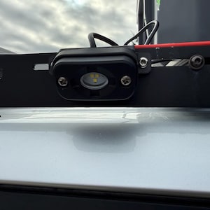 May include: A black LED light fixture mounted on a black metal frame. The light has a clear lens and is secured with two screws. A red wire is visible, along with a black wire and a silver surface.