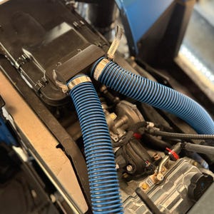 May include: Close-up of a blue engine compartment with two blue corrugated hoses connected to a black metal box. The hoses are secured with silver clamps. The engine compartment is filled with various wires and components.