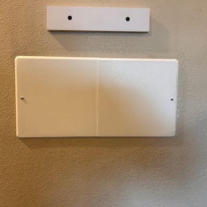 May include: A white rectangular wall plate with two screws visible on the left and right sides. The plate is mounted on a beige wall.