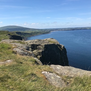Può includere: Una vista panoramica di una costa rocciosa con una scogliera erbosa che domina un'ampia distesa d'acqua blu. Il cielo è di un azzurro limpido con alcune nuvole bianche.