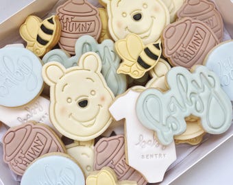 May include: A box of decorated sugar cookies featuring Winnie the Pooh, honey pots, bees, and the word "baby". The cookies are in various shades of blue, yellow, brown, and white.