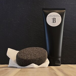 May include: A black hand and body lotion tube with the text "hand + body lotion" and "basalt B e 200ml". A dark gray pumice stone sits on a white cloth. The items are on a wooden surface with a black background.