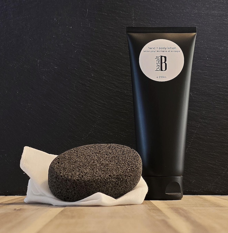 May include: A black hand and body lotion tube with the text "hand + body lotion" and "basalt B e 200ml". A dark gray pumice stone sits on a white cloth. The items are on a wooden surface with a black background.