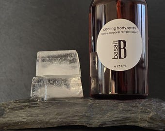 May include: A dark brown glass bottle of "cooling body spray" with a white label and the brand name "basalt". Two ice cubes sit in front of the bottle on a dark gray stone surface. The bottle contains 237 mL of product.