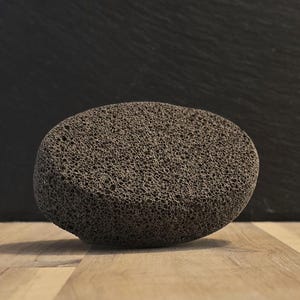 May include: A dark grey pumice stone with a porous texture. The oval-shaped stone sits on a light brown wooden surface, with a dark grey background. This natural stone is used for exfoliating skin.