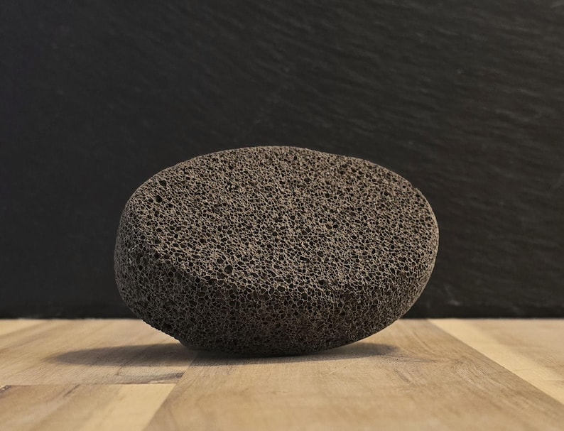 May include: A dark gray pumice stone with a porous texture. The oval-shaped stone sits on a light brown wooden surface, with a dark gray background. This natural stone is used for exfoliating skin.