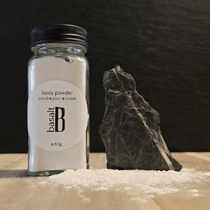 May include: A clear glass jar with a black lid contains white body powder. A white label reads "body powder, poudre pour le corps" with the brand name "basalt" and a "B" logo. A dark gray rock sits next to the jar. The jar contains 60g of powder.