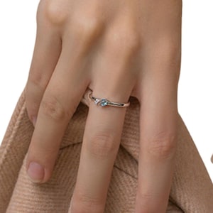 Op de afbeelding: Een zilveren ring met een kleine, ronde, lichtblauwe edelsteen. De ring zit aan een vinger, met een neutrale achtergrond. De ring heeft een delicaat ontwerp, met aan weerszijden van de belangrijkste edelsteen een kleine accentsteen.