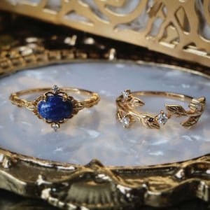 Może przedstawiać: Dwa złote pierścionki na białej i złotej ozdobnej tacy. Jeden pierścionek ma niebieski kamień szlachetny z złotymi akcentami. Drugi pierścionek to delikatny złoty wzór liści z małymi przezroczystymi kamieniami.