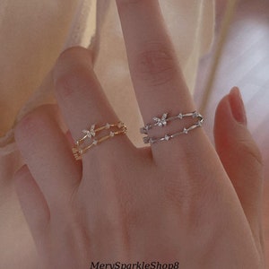 Könnte beinhalten: Zwei zarte Ringe mit einem Schmetterlingsdesign und kleinen klaren Steinen. Der Ring links ist golden und der Ring rechts ist silbern.