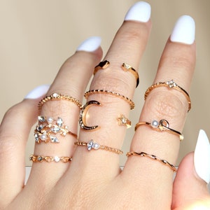 Könnte beinhalten: Ein Set goldfarbener Ringe mit verschiedenen Designs, darunter ein Halbmond, ein Stern, ein Herz und ein zartes Blumenmuster. Die Ringe sind auf einer Hand gestapelt und zeigen die filigranen Details jedes Stücks.