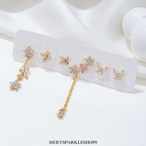 Peut inclure: Un ensemble de six boucles d'oreilles dorées avec des accents de cristal. Les boucles d'oreilles présentent une variété de motifs, notamment des fleurs, des papillons et une chaîne pendante.