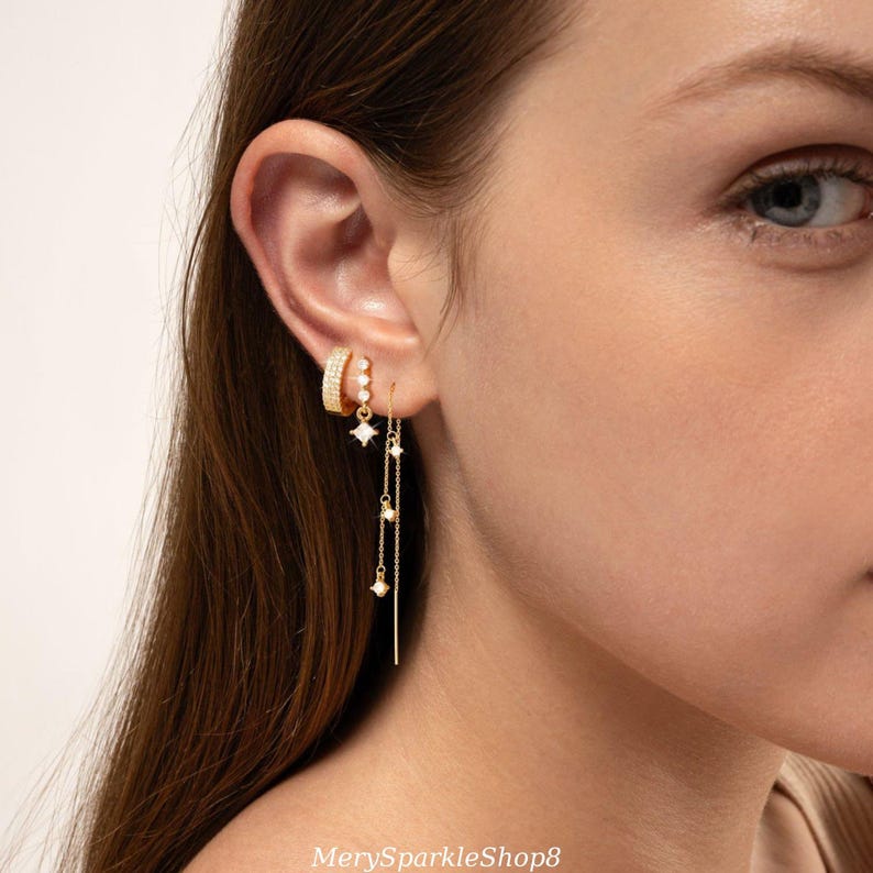 May include: A close-up of a person's ear wearing multiple gold earrings. The earrings feature small, sparkling stones and a long, delicate chain with small, dangling stones.