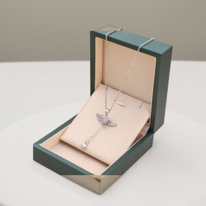 Puede incluir: Un collar de plata con un colgante de coraz&oacute;n rosa y alas de cristal transparente. El collar se muestra en una caja de joyer&iacute;a de terciopelo verde.