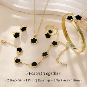 May include: A 5-piece gold jewelry set with black flower accents. The set includes two bracelets, a pair of earrings, a necklace, and a ring.