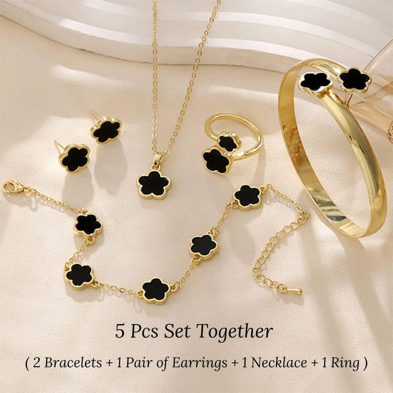 May include: A 5-piece gold jewelry set with black flower accents. The set includes two bracelets, a pair of earrings, a necklace, and a ring.