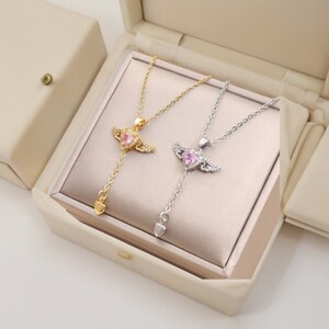 Puede incluir: Dos delicados collares con colgantes de coraz&oacute;n rosa y alas de &aacute;ngel. Los collares se muestran en una caja blanca con interior dorado.