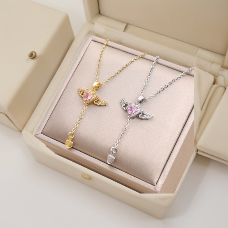 Puede incluir: Dos delicados collares con colgantes de coraz&oacute;n rosa y alas de &aacute;ngel. Los collares se muestran en una caja blanca con interior dorado.