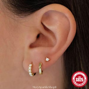 Può includere: Primo piano di un orecchio con tre orecchini: un piccolo cerchio d'oro con pietre bianche, un cerchio d'oro semplice e un orecchino a bottone d'oro con una pietra bianca. L'immagine include anche un timbro 'S925'.