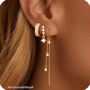 May include: Gold earrings with a hoop and dangling charms. The charms are small squares with clear stones and a long, thin chain with a small clear stone at the end.