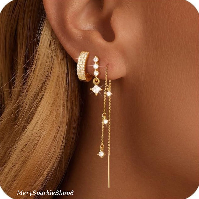 May include: Gold earrings with a hoop and dangling charms. The charms are small squares with clear stones and a long, thin chain with a small clear stone at the end.