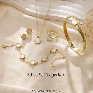 May include: A five-piece jewelry set featuring a gold chain necklace, a bracelet, a ring, and a pair of earrings. Each piece has a white flower-shaped pendant with a gold border. The text "5 Pcs Set Together" is visible below the jewelry.