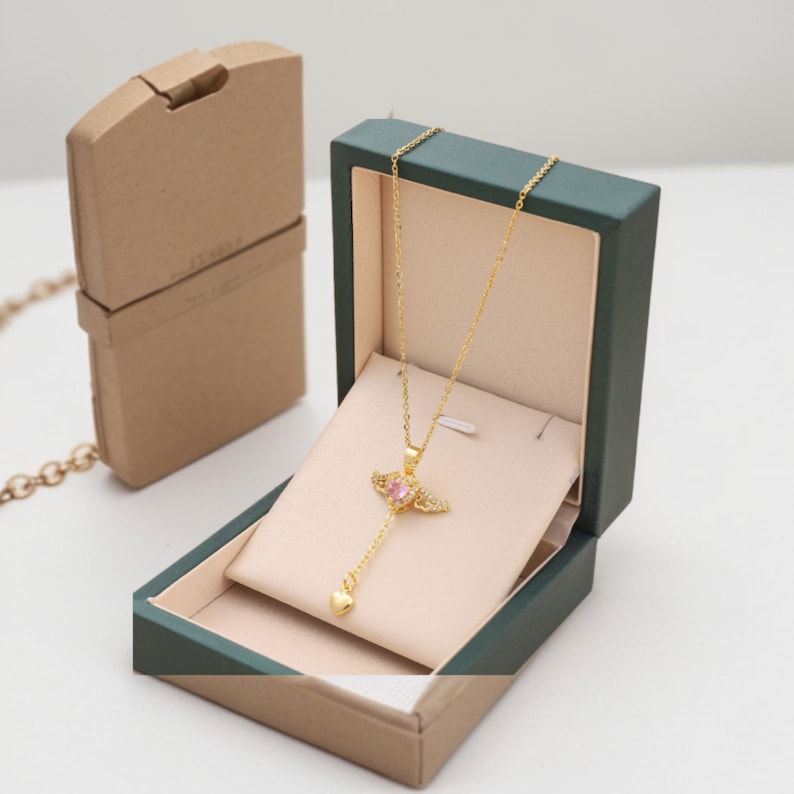Puede incluir: Un collar de oro con un colgante de cristal rosa en forma de coraz&oacute;n con alas. El collar se muestra en una caja de terciopelo verde con un interior beige.