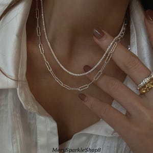 Op de afbeelding: Twee zilveren kettingen die op elkaar gelegd zijn op de nek van een persoon. De bovenste ketting is een dunne ketting met kleine, ronde stenen. De onderste ketting is een dikkere ketting met grotere, ovale schakels.