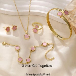May include: A five-piece jewelry set featuring a gold chain necklace, a bracelet, a ring, a pair of earrings, and a bangle bracelet. Each piece has a pink flower charm.