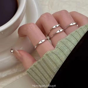May include: Four silver rings, each with a different letter: C, W, X, and Y. The rings are on fingers, with a light green sweater sleeve in the background. A cup of coffee is visible on the left.