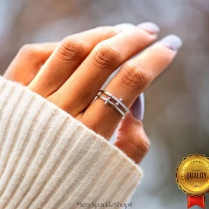 Puede incluir: Un anillo plateado con un diseño de doble banda, con un acento en forma de cruz adornado con pequeñas piedras brillantes. El anillo se lleva en un dedo, con un suéter color crema en el fondo. Es una joya.