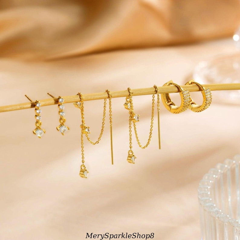 May include: A set of gold-toned earrings with clear cubic zirconia stones. The set includes a pair of hoop earrings, a pair of dangle earrings, and a pair of chain earrings.