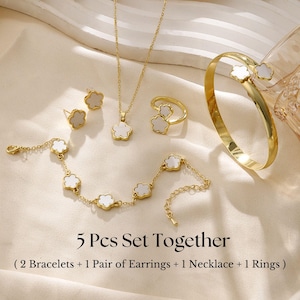 May include: A gold-toned jewelry set featuring a necklace, ring, earrings, and two bracelets. Each piece has a flower-shaped design with a white center. The text "5 Pcs Set Together" is visible.