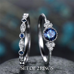 Puede incluir: Un conjunto de dos anillos de plata con piedras preciosas azules. El anillo más grande presenta una piedra preciosa azul redonda rodeada de piedras preciosas blancas más pequeñas. El anillo más pequeño tiene una fila de piedras preciosas azules y blancas.