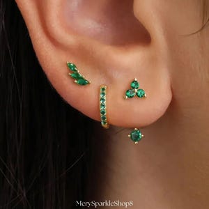 May include: A close-up of an ear adorned with multiple gold earrings featuring emerald green gemstones. The earrings include a leaf-shaped stud, a small hoop, a cluster of three stones, and a square stud. The shop name "MerySparkleShop8" is at the bottom.