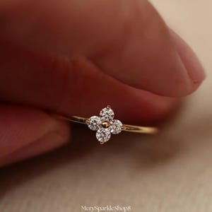 Puede incluir: Un delicado anillo de oro con un diseño de trébol de cuatro hojas. El trébol está compuesto por cuatro diamantes redondos y brillantes. El anillo está sostenido por una mano sobre un fondo suave y borroso. El nombre de la tienda "MerySparkleShop8" es visible.