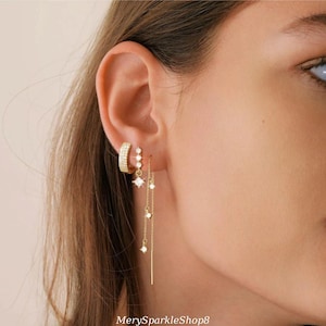 May include: A close-up of a person's ear with multiple gold earrings. The earrings feature small, clear cubic zirconia stones and a delicate chain design.