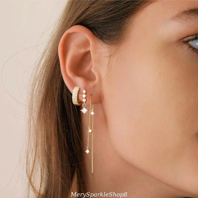 May include: A close-up of a person's ear with multiple gold earrings. The earrings feature small, clear cubic zirconia stones and a delicate chain design.