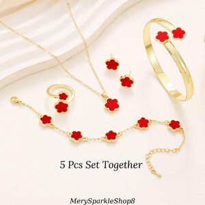 May include: A 5 piece jewelry set featuring a gold chain necklace, bracelet, ring, and earrings. Each piece has a red flower charm.