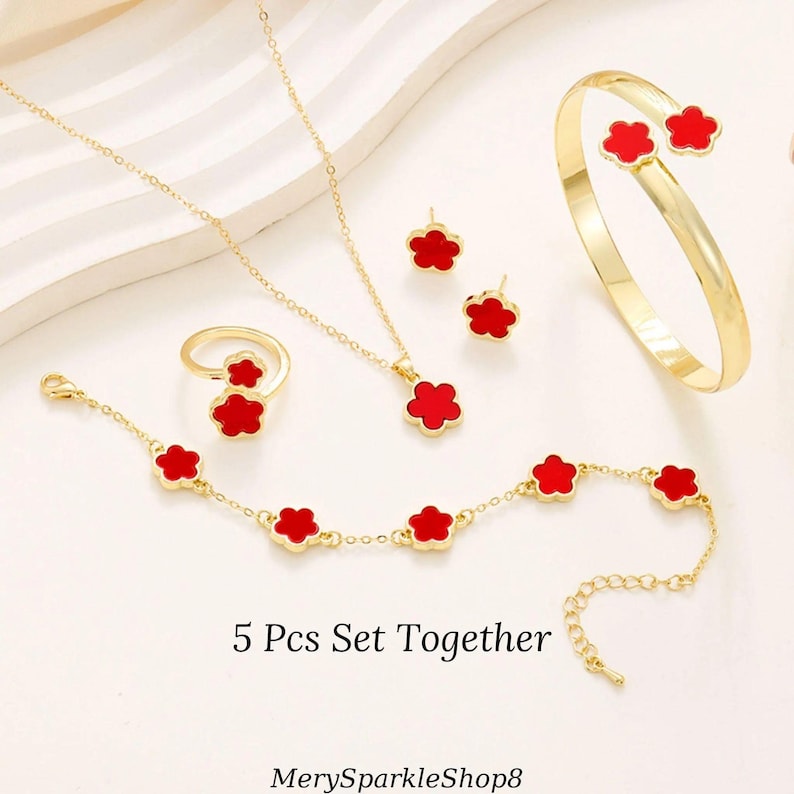 May include: A 5 piece jewelry set featuring a gold chain necklace, bracelet, ring, and earrings. Each piece has a red flower charm.