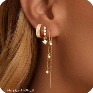 May include: Gold earrings with a hoop and a dangling chain with small cubic zirconia stones. The chain has a square cubic zirconia stone at the top and a smaller stone at the bottom.
