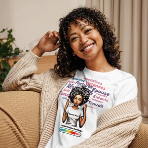 May include: White t-shirt featuring a colourful graphic of a woman with a thought bubble filled with words such as "vacances", "factures", "épouse", and "enfants". The graphic also includes a progress bar labelled "Charge mentale Warning... 90%".