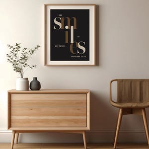 Könnte beinhalten: Ein gerahmter Wandkunstdruck mit schwarzem Hintergrund und dem weißen sowie beigefarbenen Text "SHE SMILES AT HER FUTURE PROVERBS 31:25". Die Typografie ist geschichtet und stilisiert, wobei das Wort "SMILES" im Mittelpunkt steht.