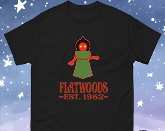 May include: Black t-shirt with a red and green illustration of a humanoid figure with glowing red eyes. The text "FLATWOODS -EST. 1952- " is printed below the figure.