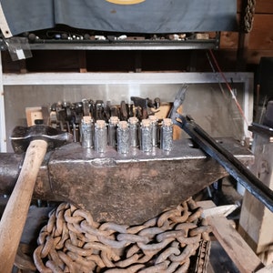 May include: A close-up of a metal anvil with a chain wrapped around it. There are 10 small glass vials with cork tops sitting on top of the anvil. The anvil is in a workshop setting with tools and a Minnesota state flag hanging in the background.