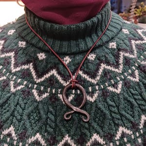 May include: A handcrafted, dark metal pendant necklace. The pendant is an abstract shape suspended from a brown leather cord. The background features a green and white patterned sweater.