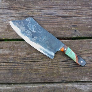 May include: A large, rustic cleaver with a dark, textured blade and a colorful handle. The handle features shades of green, orange, and brown. The cleaver rests on a weathered wooden surface.