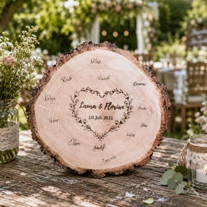 May include: A rustic wooden wedding sign with a heart-shaped floral design and the names of guests. The sign reads "Laura & Florian 10. Juli.2021". The wooden sign is placed on a wooden table with a blurred background of a wedding reception.